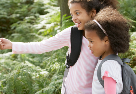 two girls with a backpack in a forest with the taller girl pointing at something 