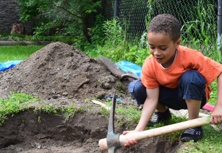 A boy in orange t-shirt digging in the dirt with a pickaxe