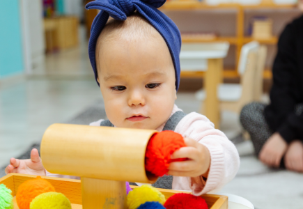 little kid with a blue headband putting an orange pompom into a small wooden cylindrical structure. 