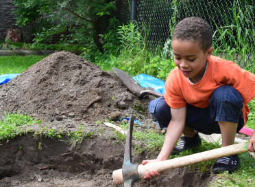 A boy in orange t-shirt digging in the dirt with a pickaxe