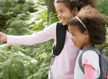 two girls with a backpack in a forest with the taller girl pointing at something 