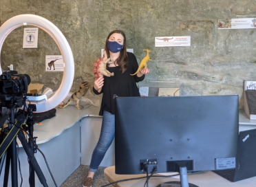 woman in mask holding dinosaur toys with ring light and computer setup in front