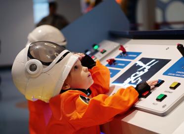 Child wearing astronaut costume lifts helmet slightly over head to see